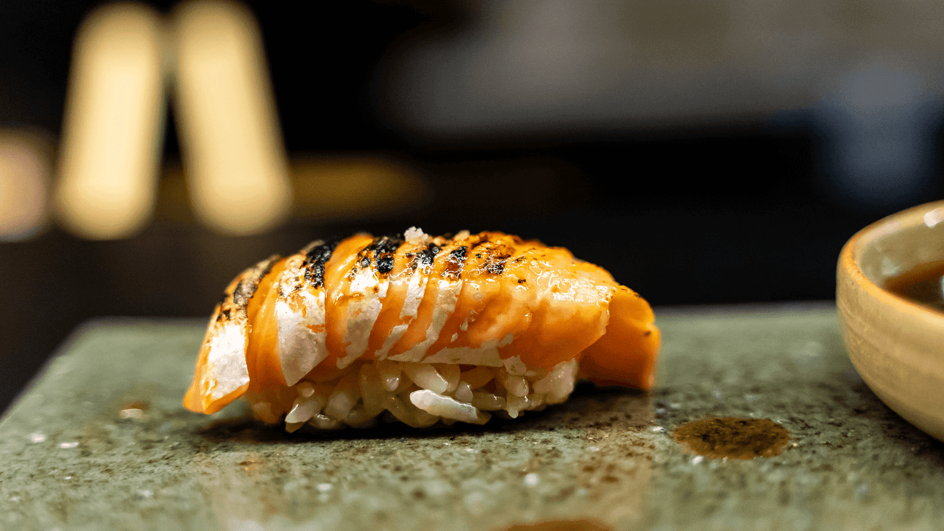 Close-up of a grilled salmon sushi piece on a green plate, with a bowl of soy sauce on the side.