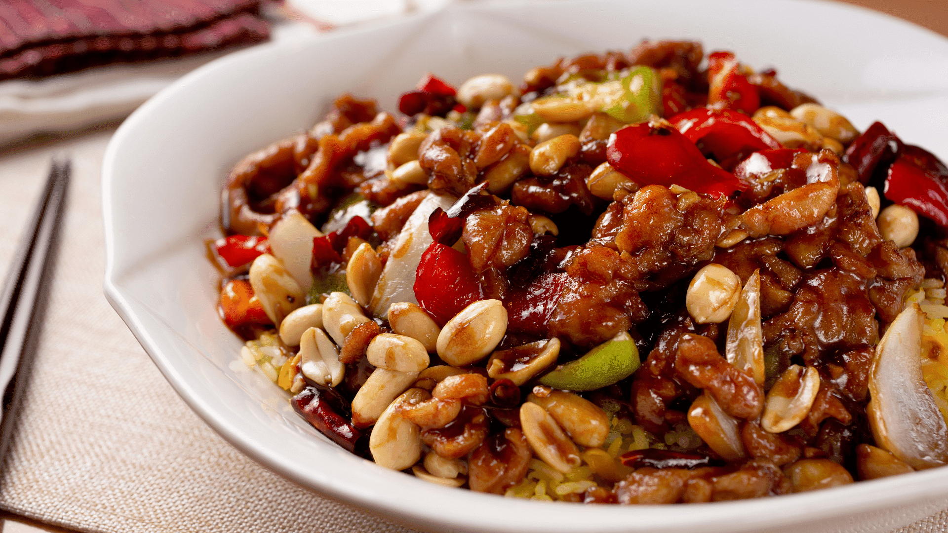 Plate of Kung Pao chicken with peanuts, bell peppers, and chili peppers on a white tablecloth.