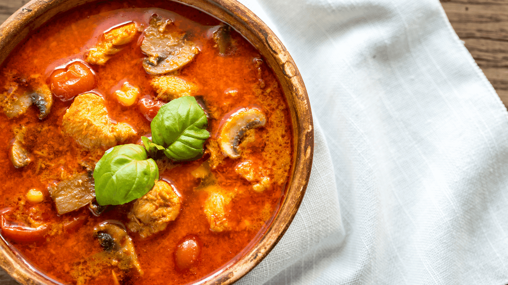 Bowl of tomato-based chicken and mushroom soup garnished with basil leaves, placed on a napkin.