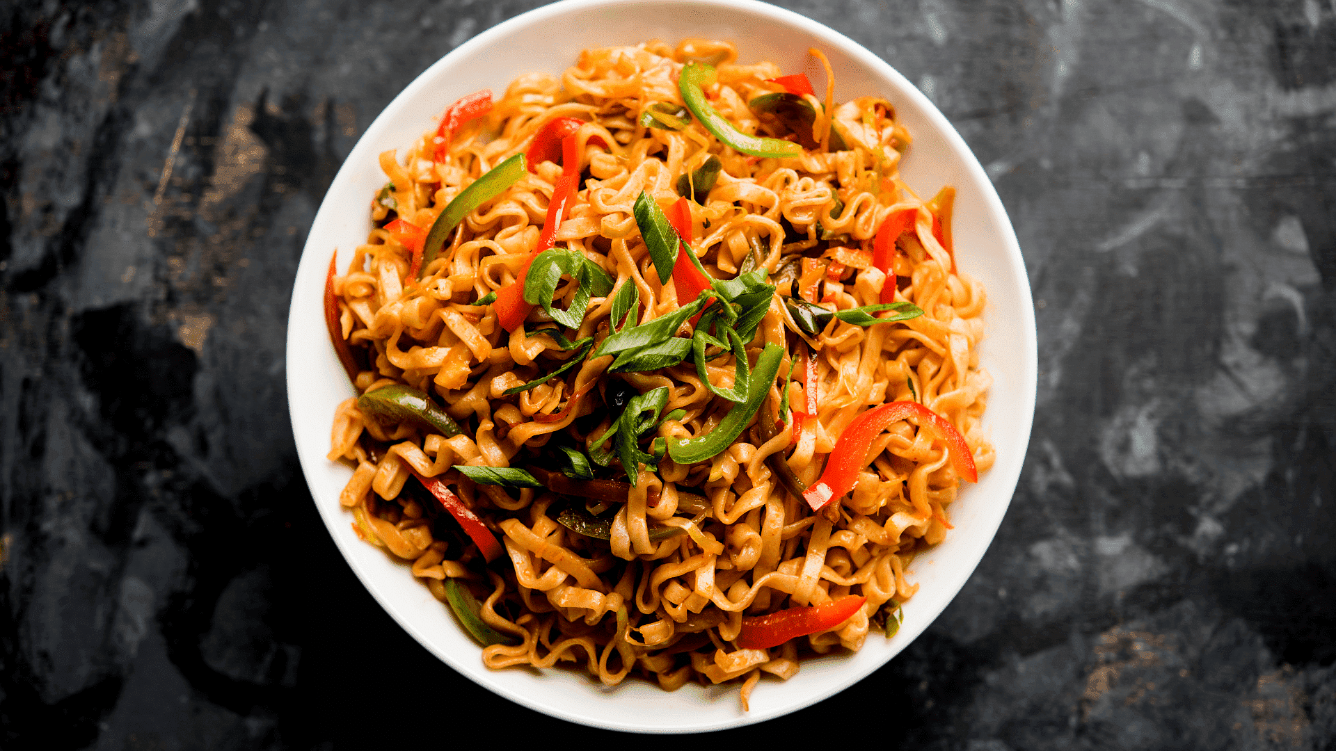 Bowl of stir-fried noodles with bell peppers and scallions on a rustic dark background.