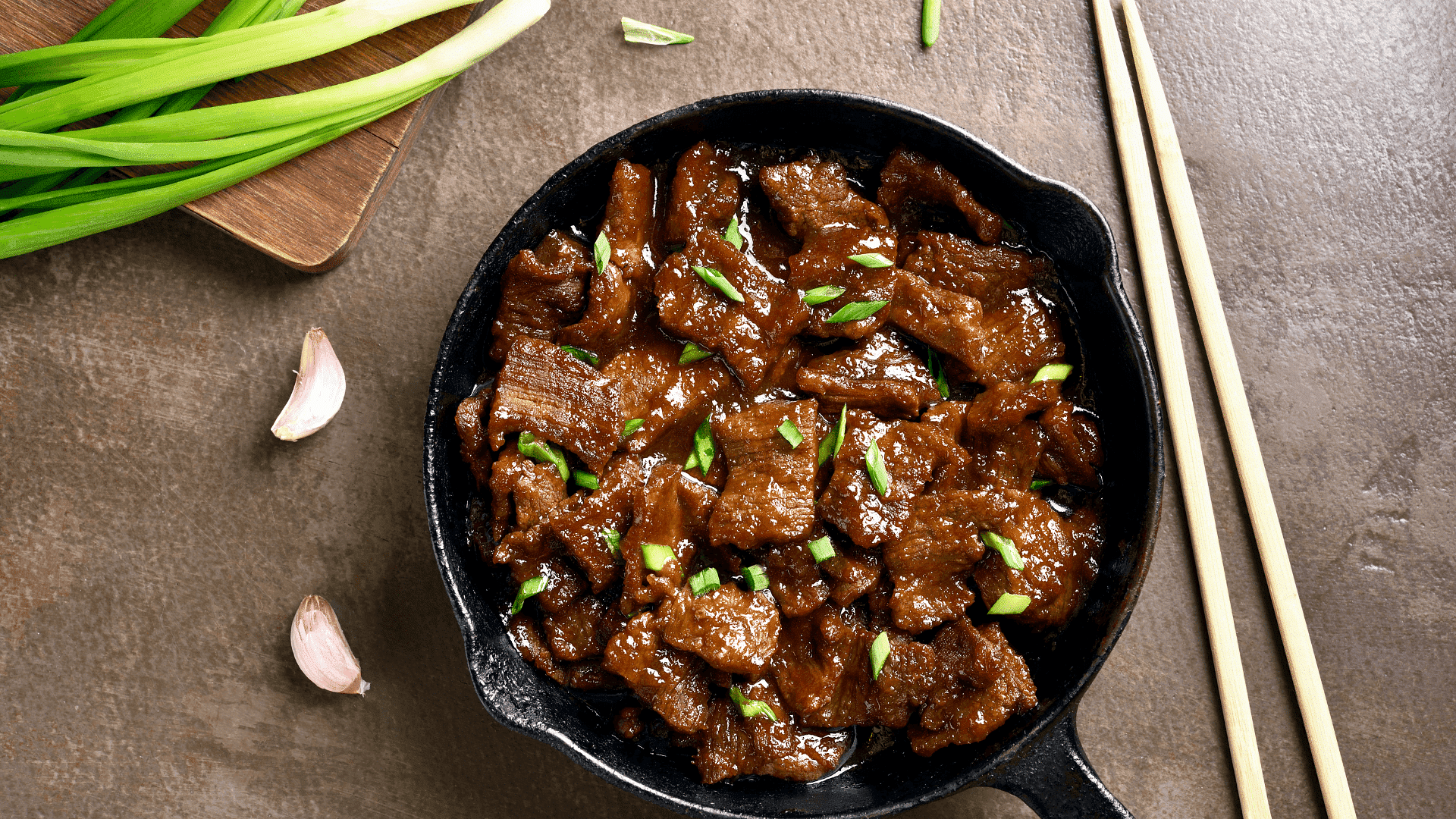 Stir-fried beef topped with green onions in a cast iron skillet next to garlic and green onions.