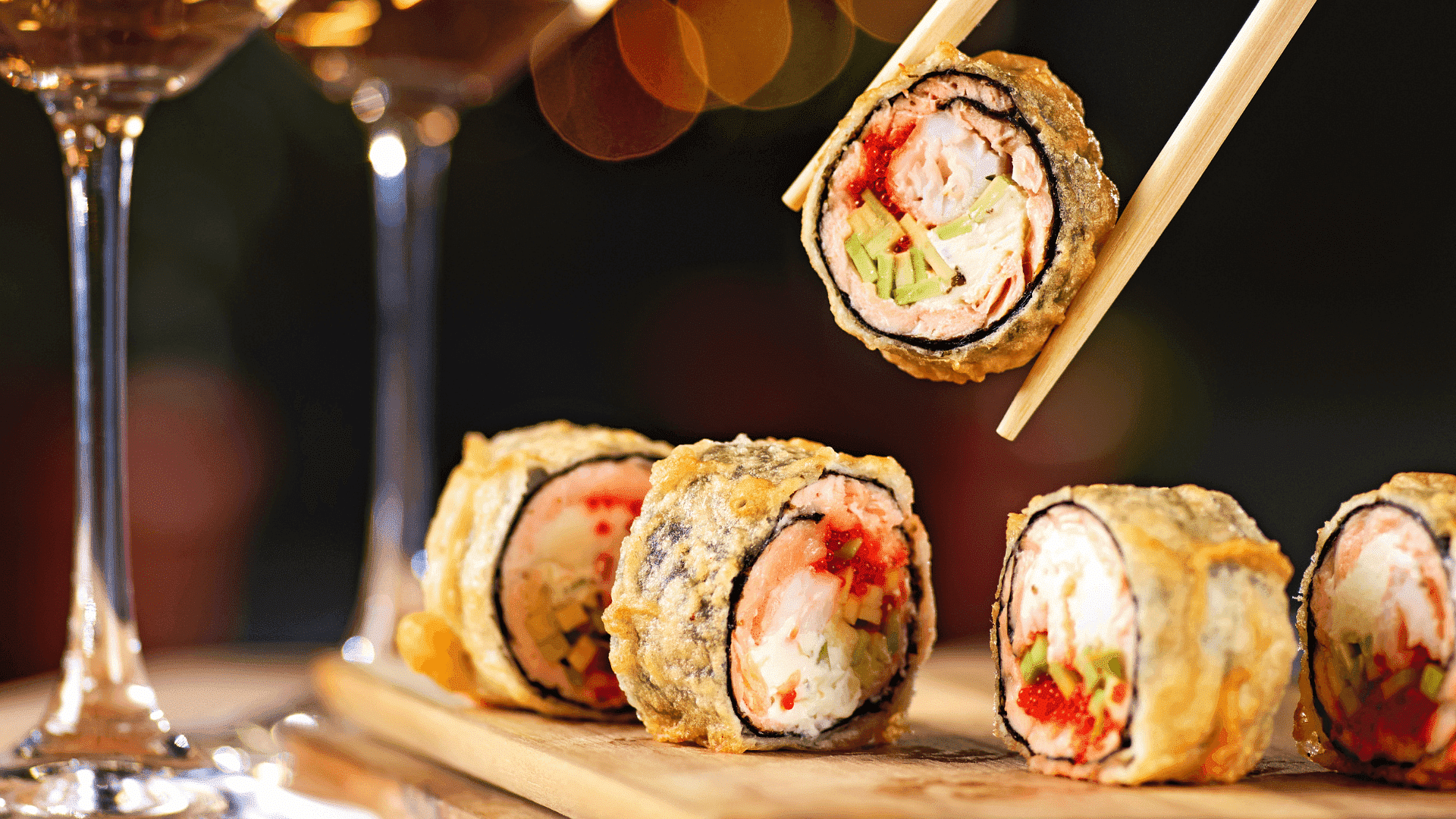 Five pieces of tempura sushi on a wooden board, with chopsticks lifting one piece.