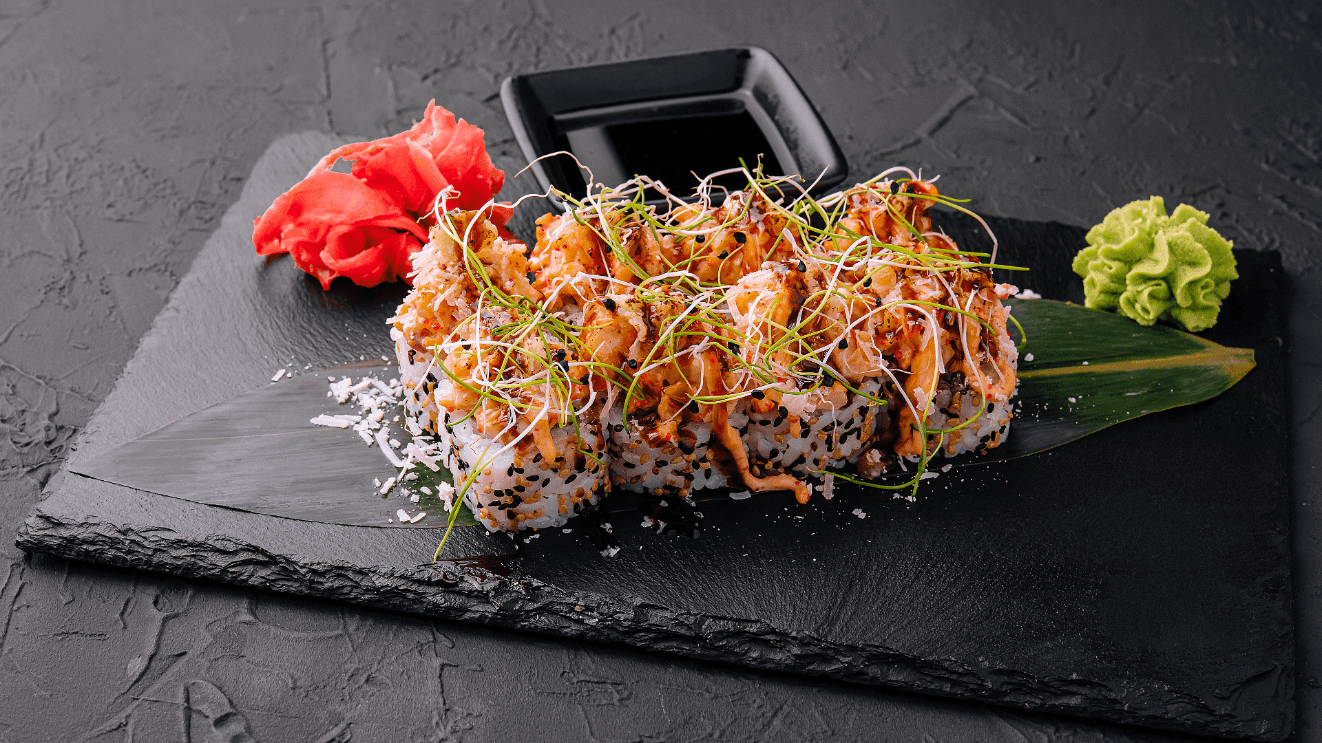 Sushi roll topped with sprouts and sauce, served with pickled ginger and wasabi on a slate plate.