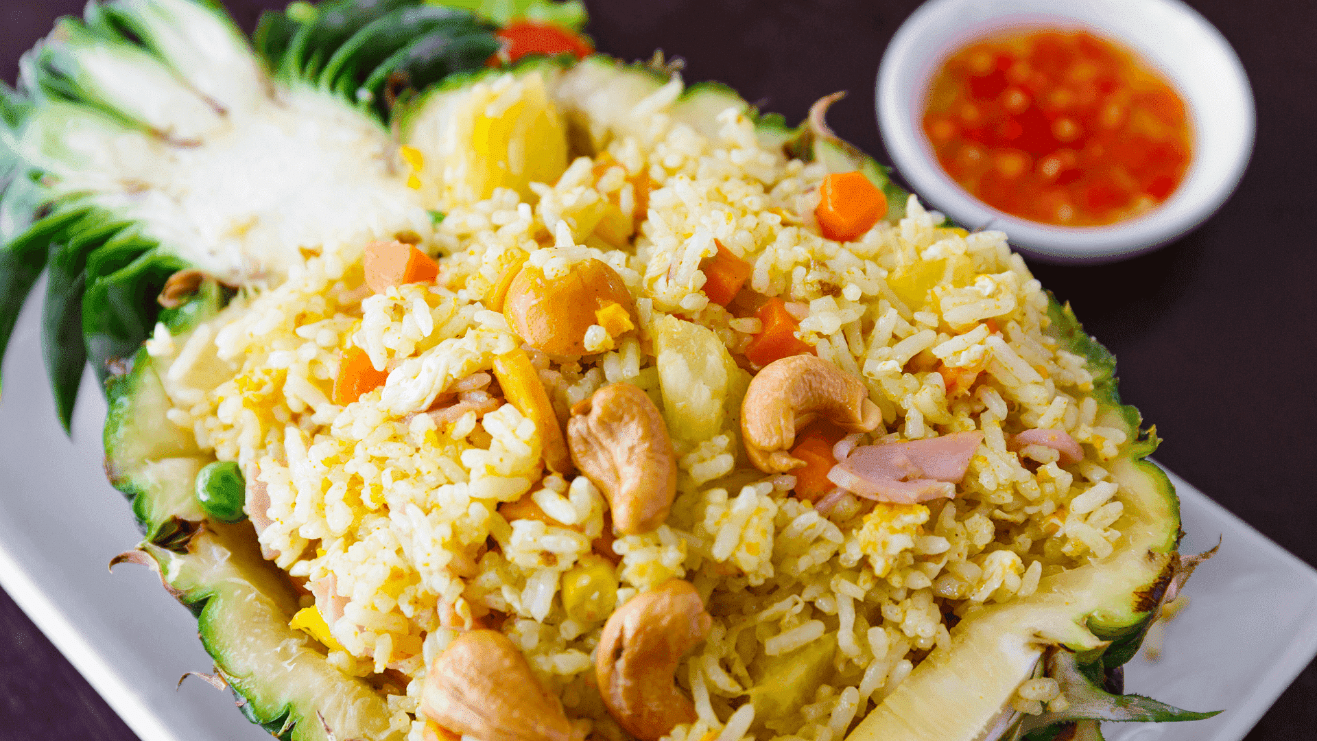 Pineapple fried rice served in a pineapple shell with cashews and a side of chili sauce.