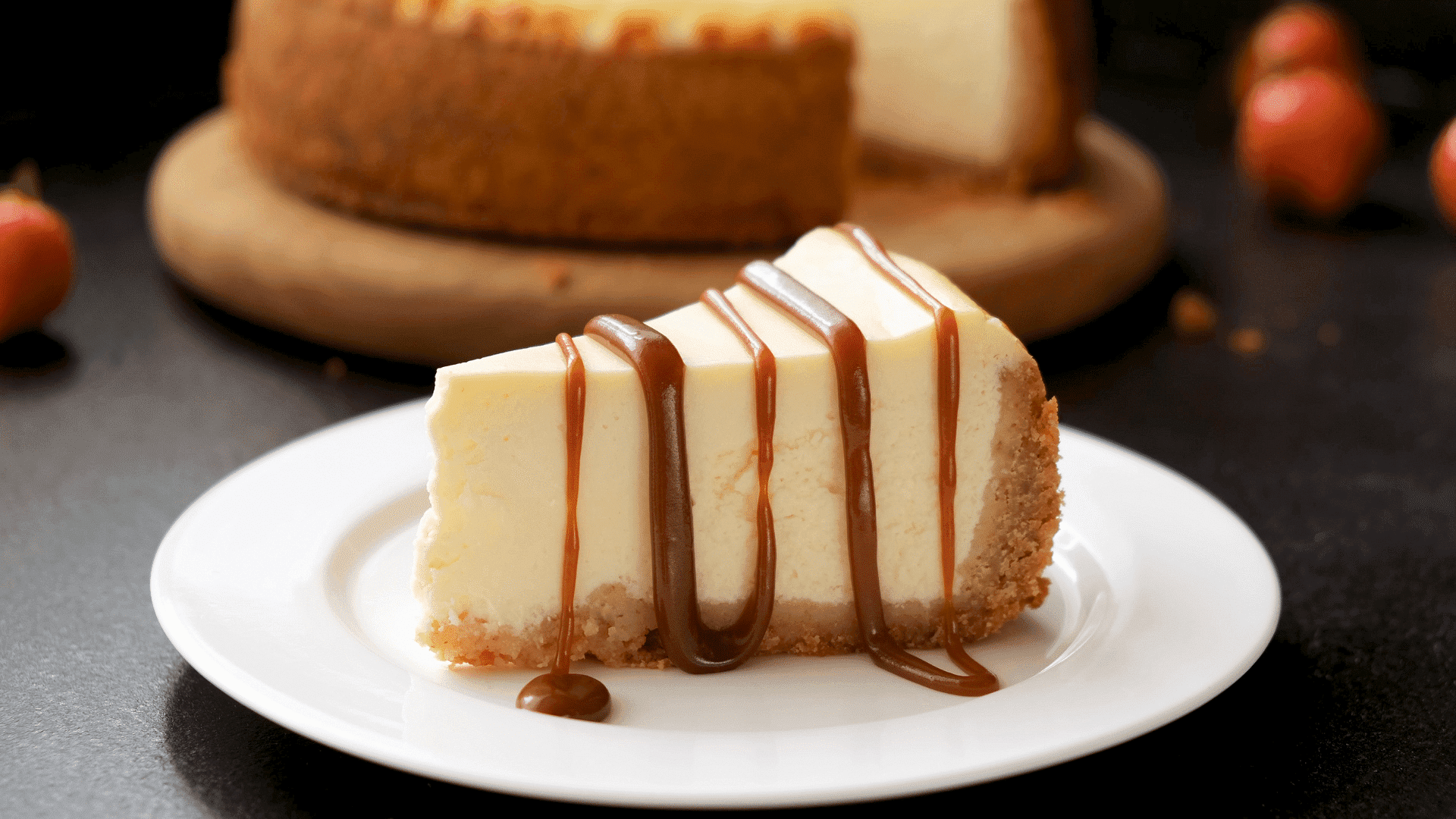 A slice of cheesecake drizzled with caramel sauce on a white plate, with whole cake in background.