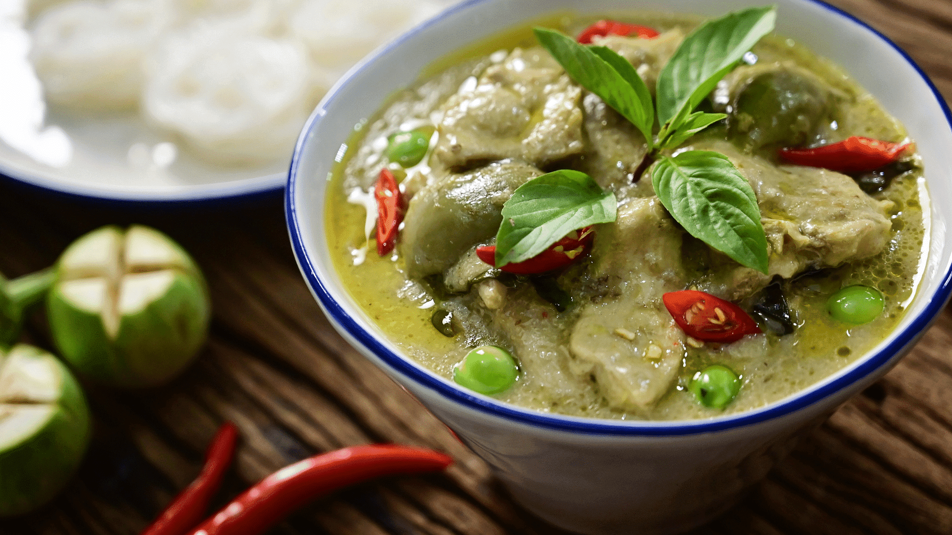 Green curry with herbs, chilies, and vegetables in a bowl, served beside a plate of rice noodles.